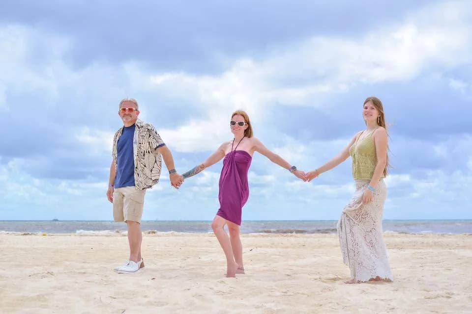 Jamie, Bob and Aubree holding hands on a wide sandy beach under a cloudy sky, smiling toward the camera.